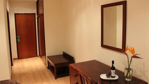 A hotel room with a wooden desk, a mirror, a small table with a bottle and glass, and a door leading to another room.