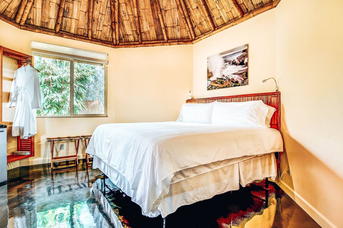 A room with a bed, a window, a painting, and a wooden ceiling.