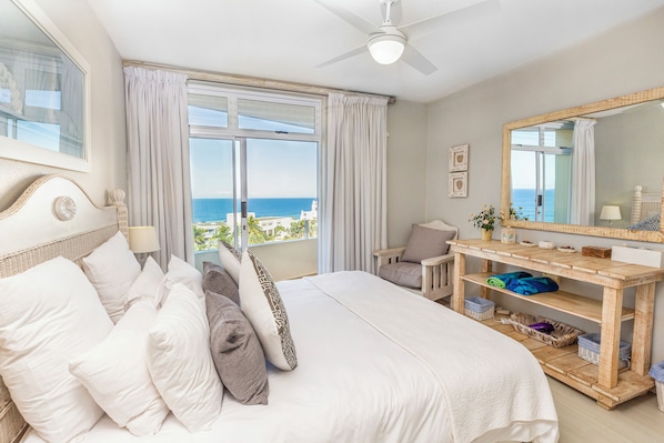 A bedroom with a bed, a vanity with a mirror, a window with curtains, and a view of the sea.