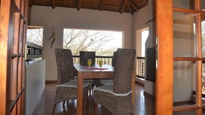A dining area with a wooden table and woven chairs, a thatched roof, and a view of trees outside. - Thabaledi Game Lodge (Beestekraal)