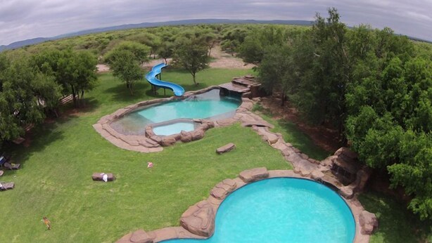 A large outdoor swimming pool with a slide, surrounded by lush greenery and a clear sky. - Thabaledi Game Lodge (Beestekraal)
