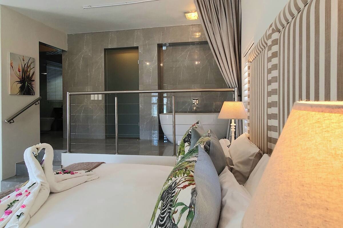 Ein modernes Hotelzimmer mit Balkon, einem großen Bett mit floralen Kissen sowie Blick auf den Pool und einem gestreiften Kopfteil.