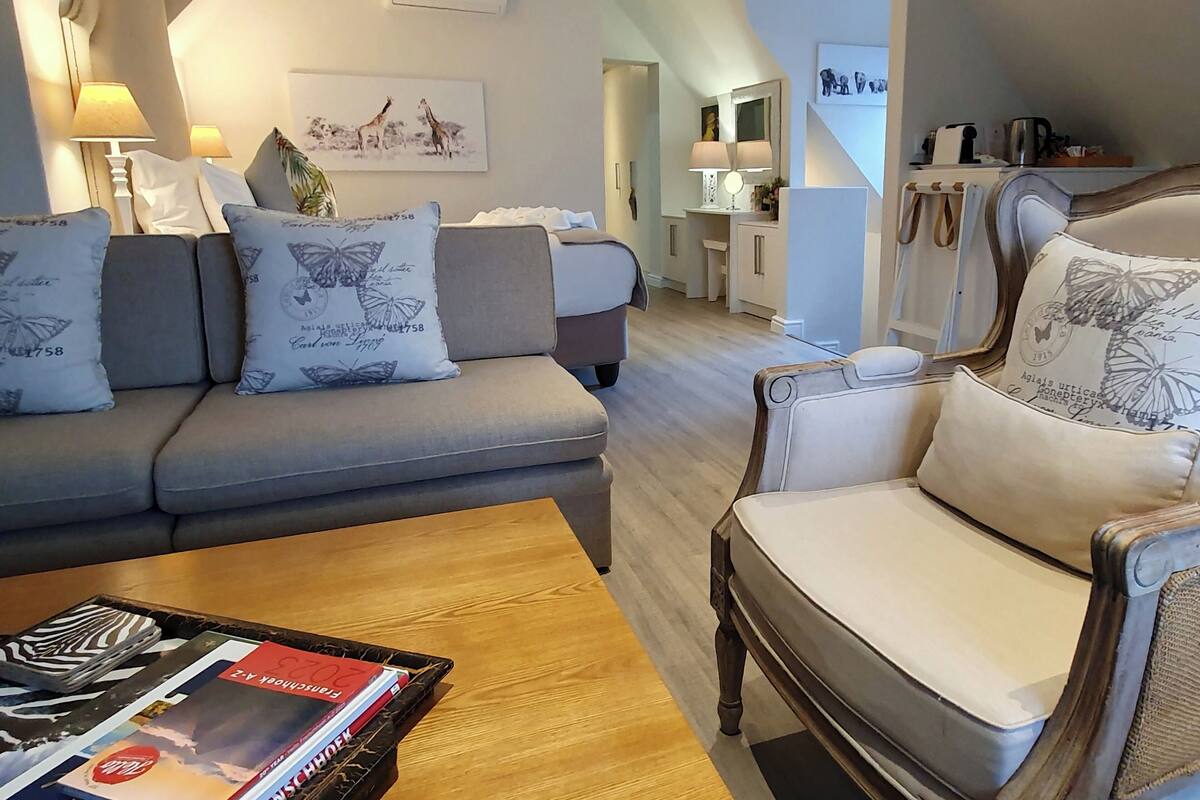 Ein modernes Wohnzimmer mit einem grauen Sofa, einem Holztisch und einem weißen Sessel. Im Hintergrund befindet sich eine Küchenzeile mit einem Esstisch und Stühlen.