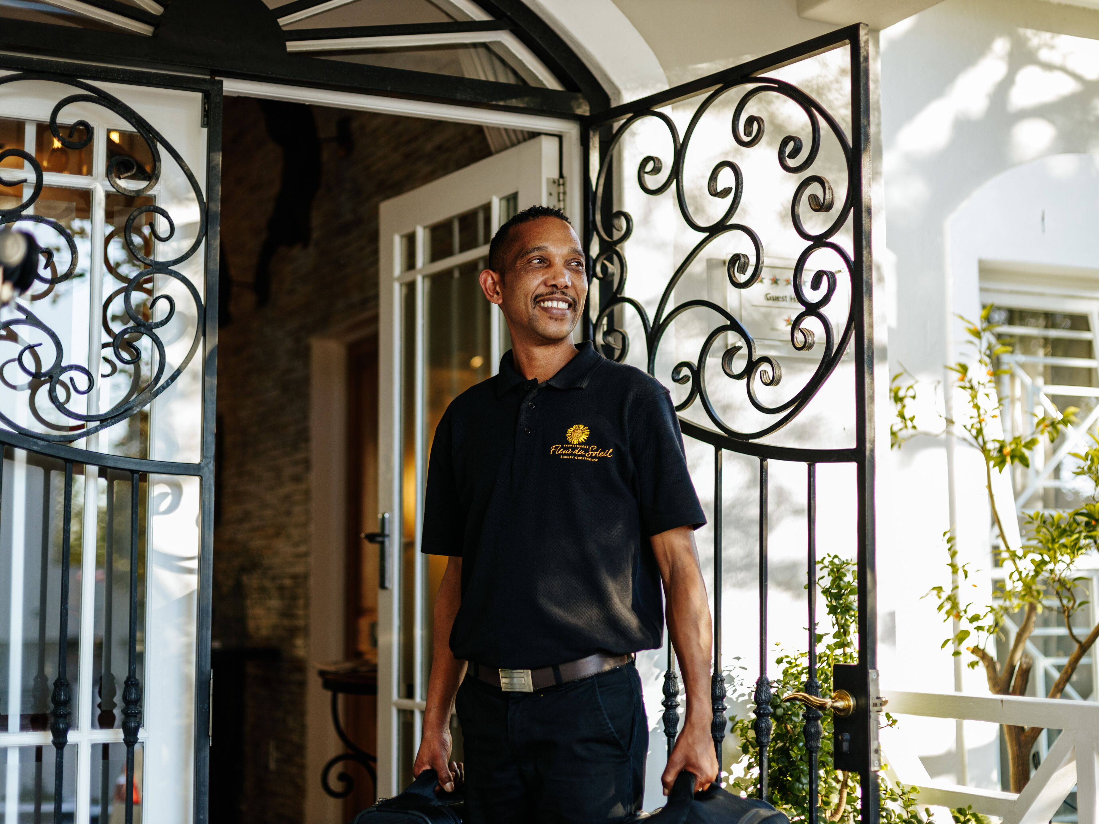 A man standing at a hotel entrance with a decorative wrought iron gate, a brick wall, and a sign indicating 'Guest House'.
