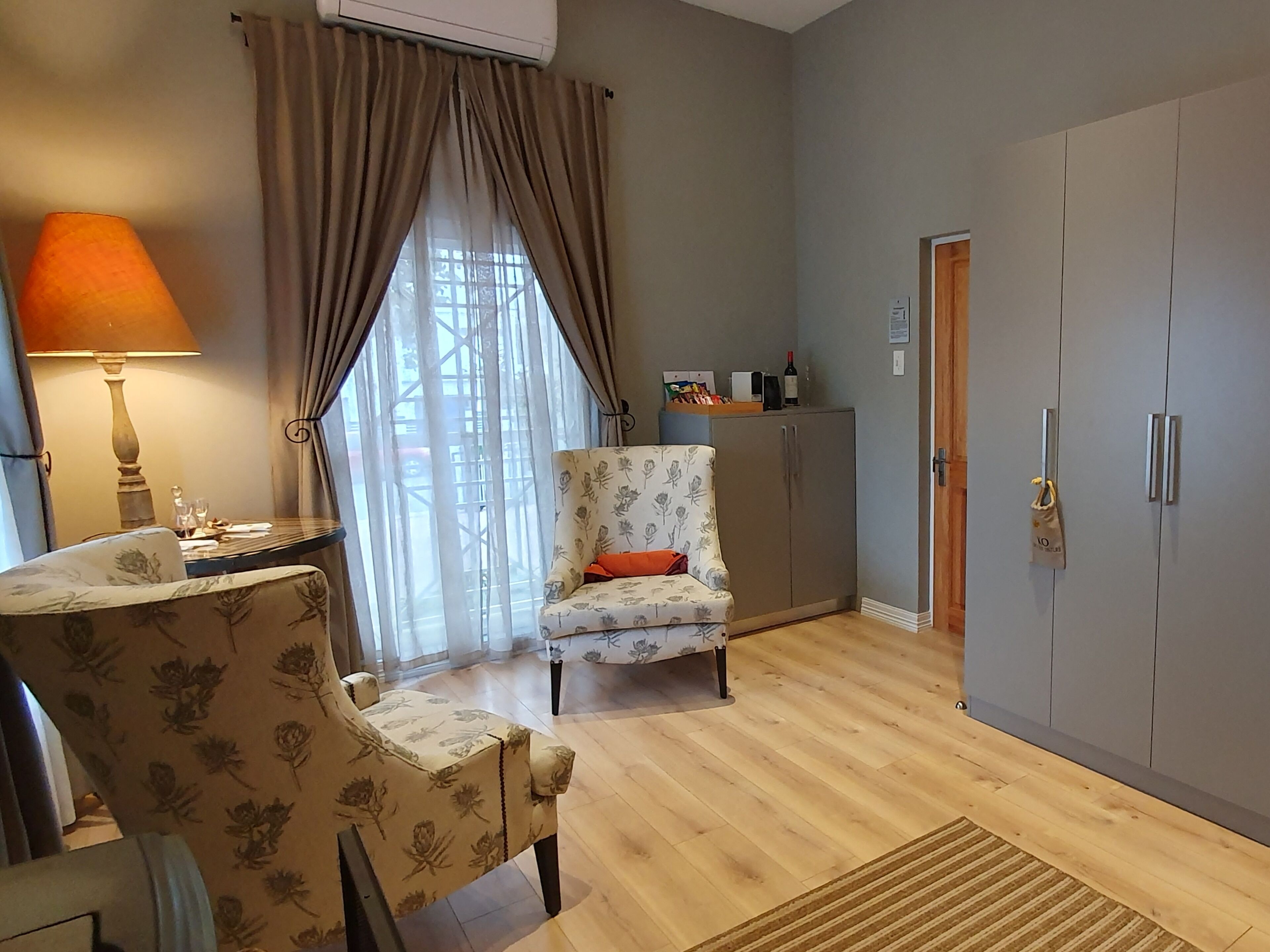 A room with a wooden floor, a floral armchair, a table with a lamp, a wardrobe, and a window with curtains.
