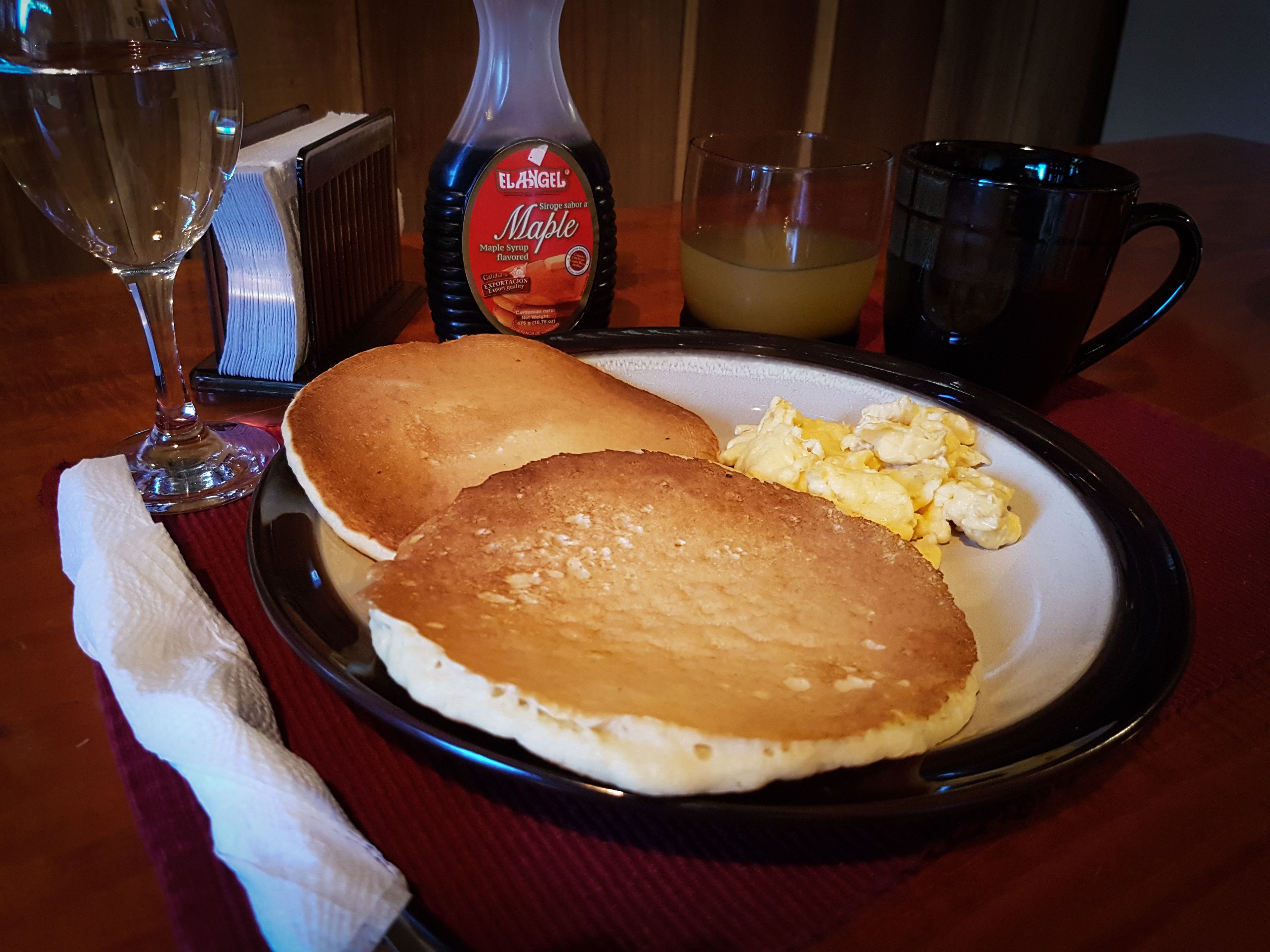 A breakfast plate with pancakes, scrambled eggs, a bottle of maple syrup, a glass of orange juice, and a coffee mug on a wooden table.