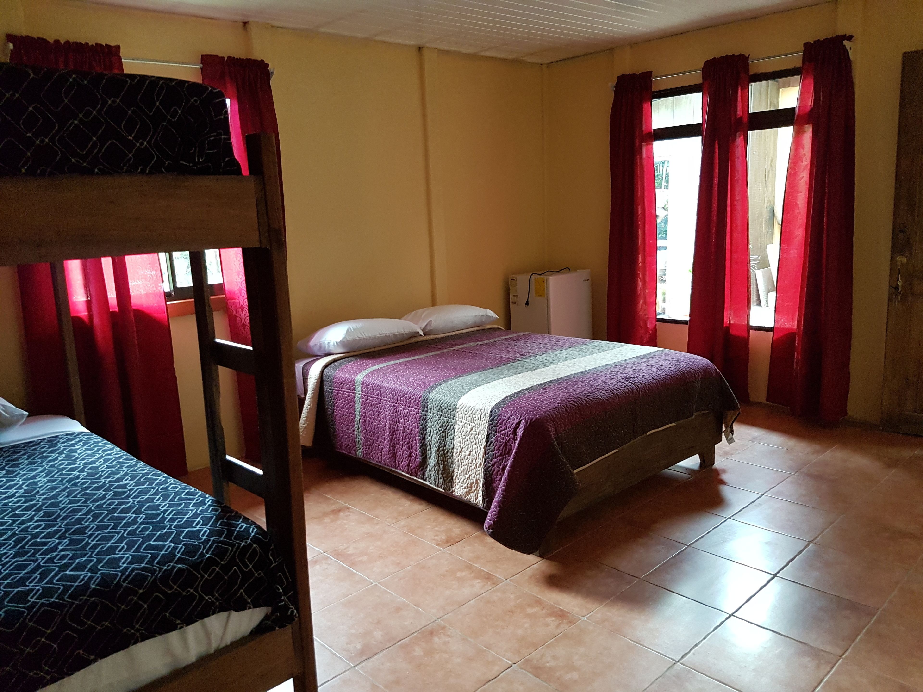 A room with a bunk bed, a single bed, a radiator, and a window with red curtains.