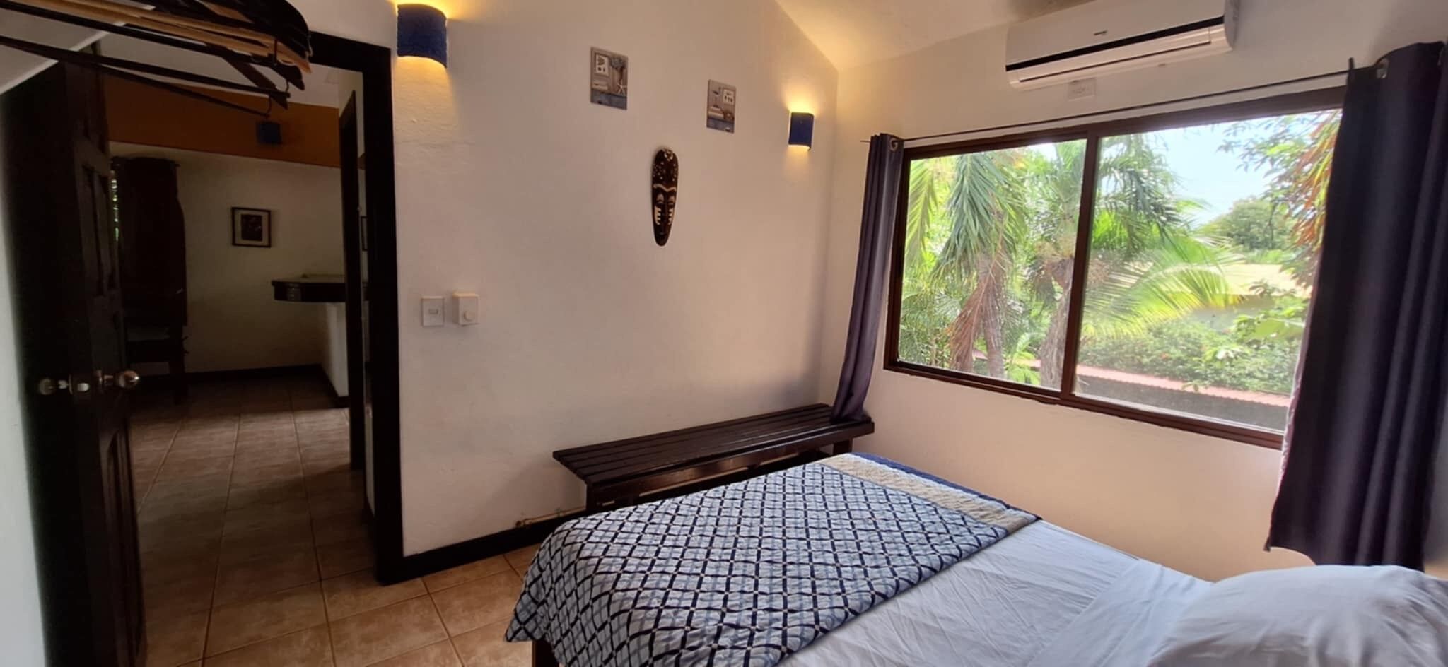 Un dormitorio con cama, ventana con vista a follaje, banca y puerta que da a otra habitación.