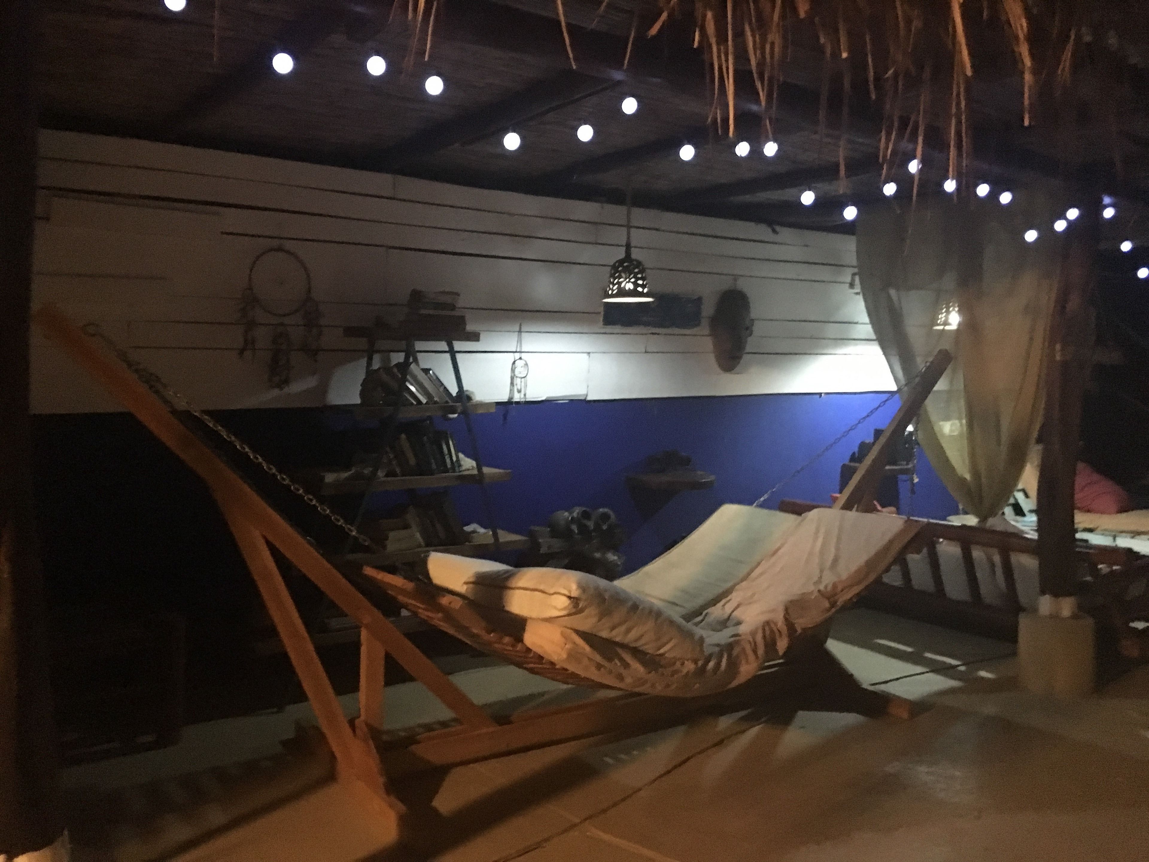 A hammock chair under a thatched roof, a shelf with various items, and a blue wall with a dreamcatcher.