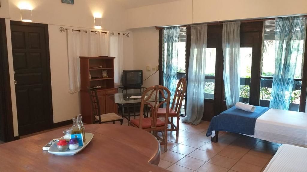 A room with a wooden dining table, a TV, a bookshelf, two single beds, and a balcony with curtains.