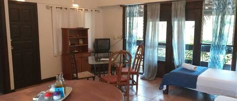 A room with a wooden dining table, a TV, a bookshelf, two single beds, and a balcony with curtains.