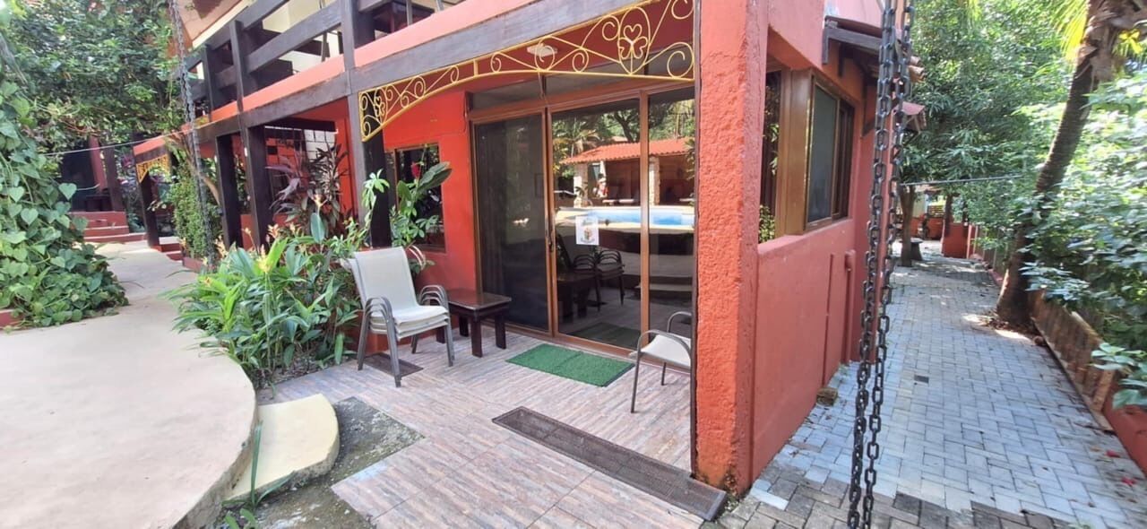 A building with a red exterior, a covered patio area with chairs and a table, and a pool visible through the glass doors.