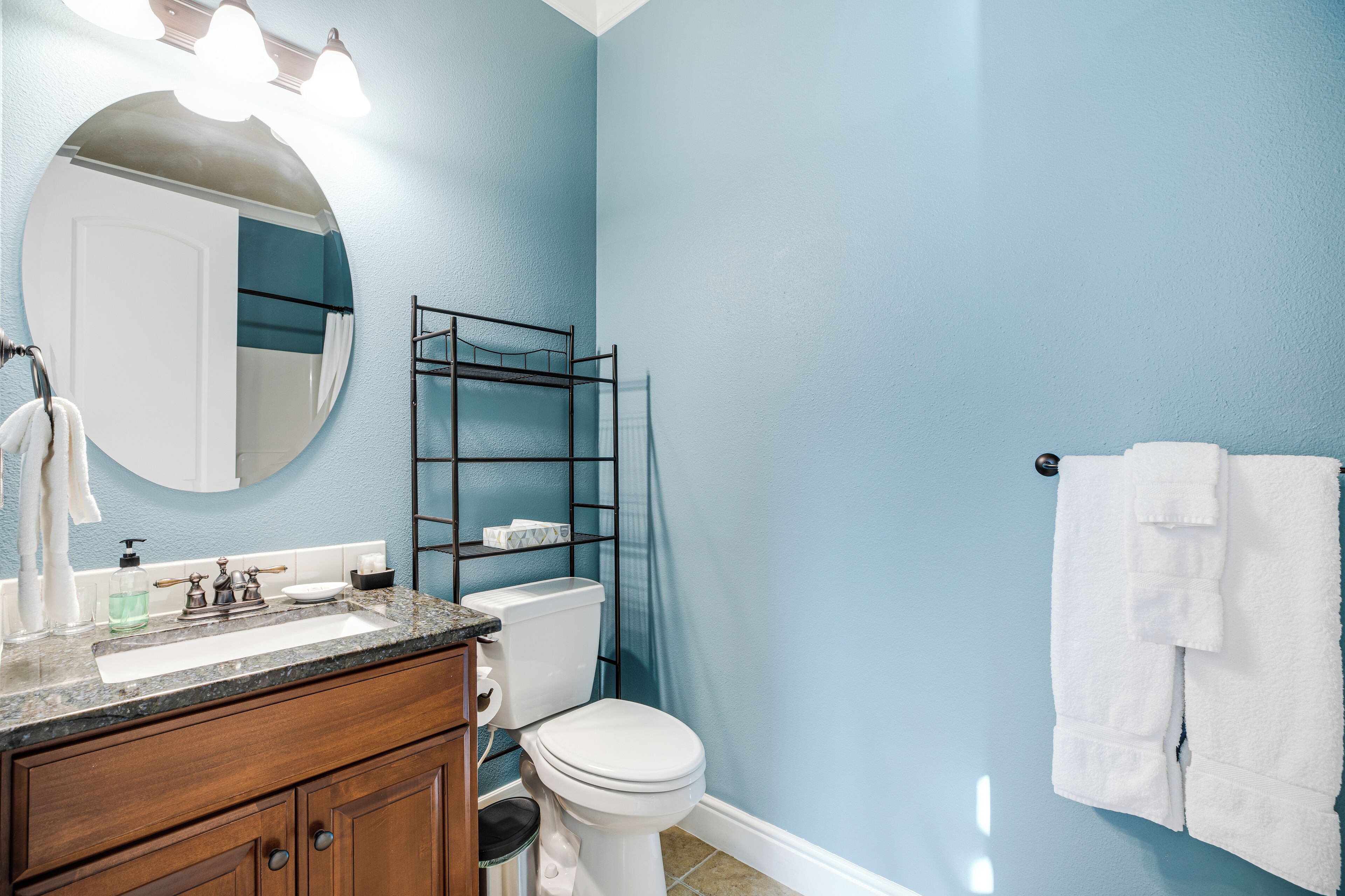 Un baño con pared azul, inodoro blanco, tocador de madera con lavamanos y un perchero para toallas.