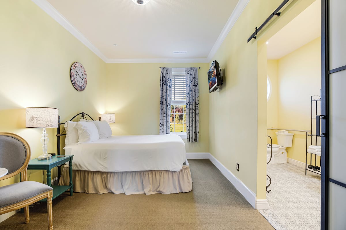 A bedroom with a bed, a chair, a table, a lamp, a clock, a window with curtains, a television, and a bathroom with a toilet and sink.
