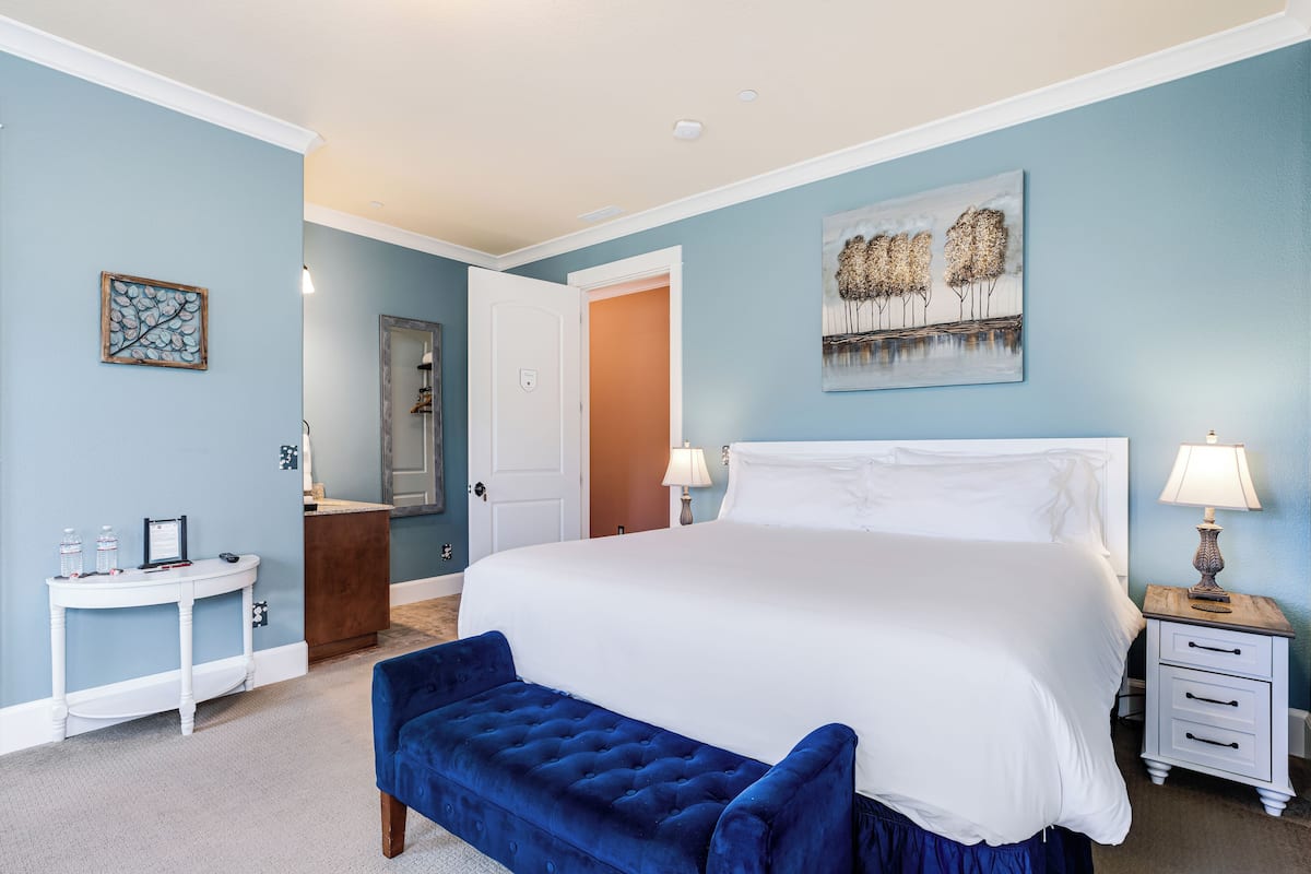 A bedroom with a large bed, a bedside table, a bench, a nightstand, and a painting on the wall.