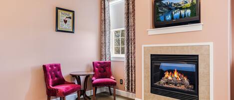 A living room with a fireplace, two red velvet chairs, a small table, and a television mounted above the fireplace.