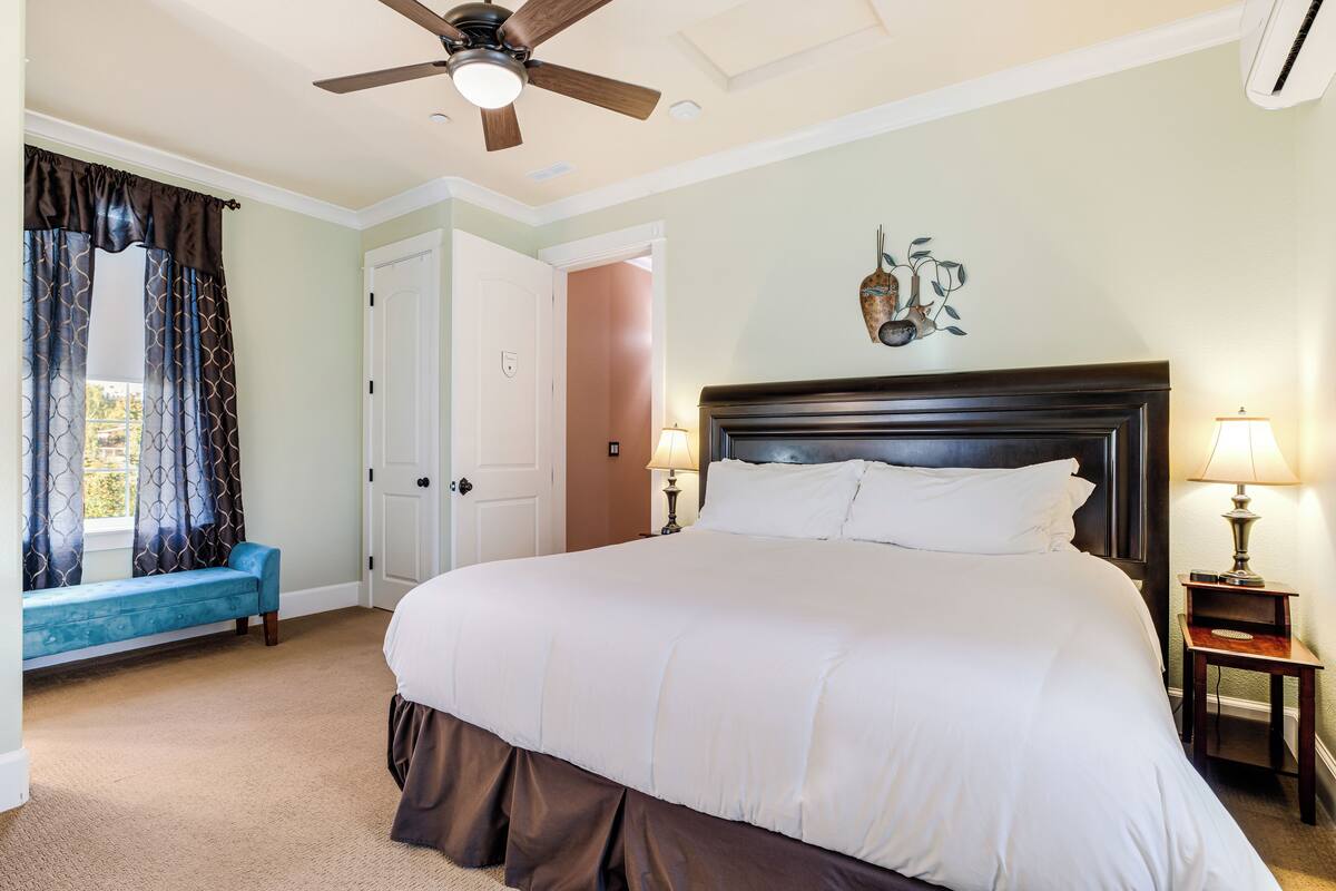 A bedroom with a large bed, a ceiling fan, a bench, a door, and a wall-mounted lamp.