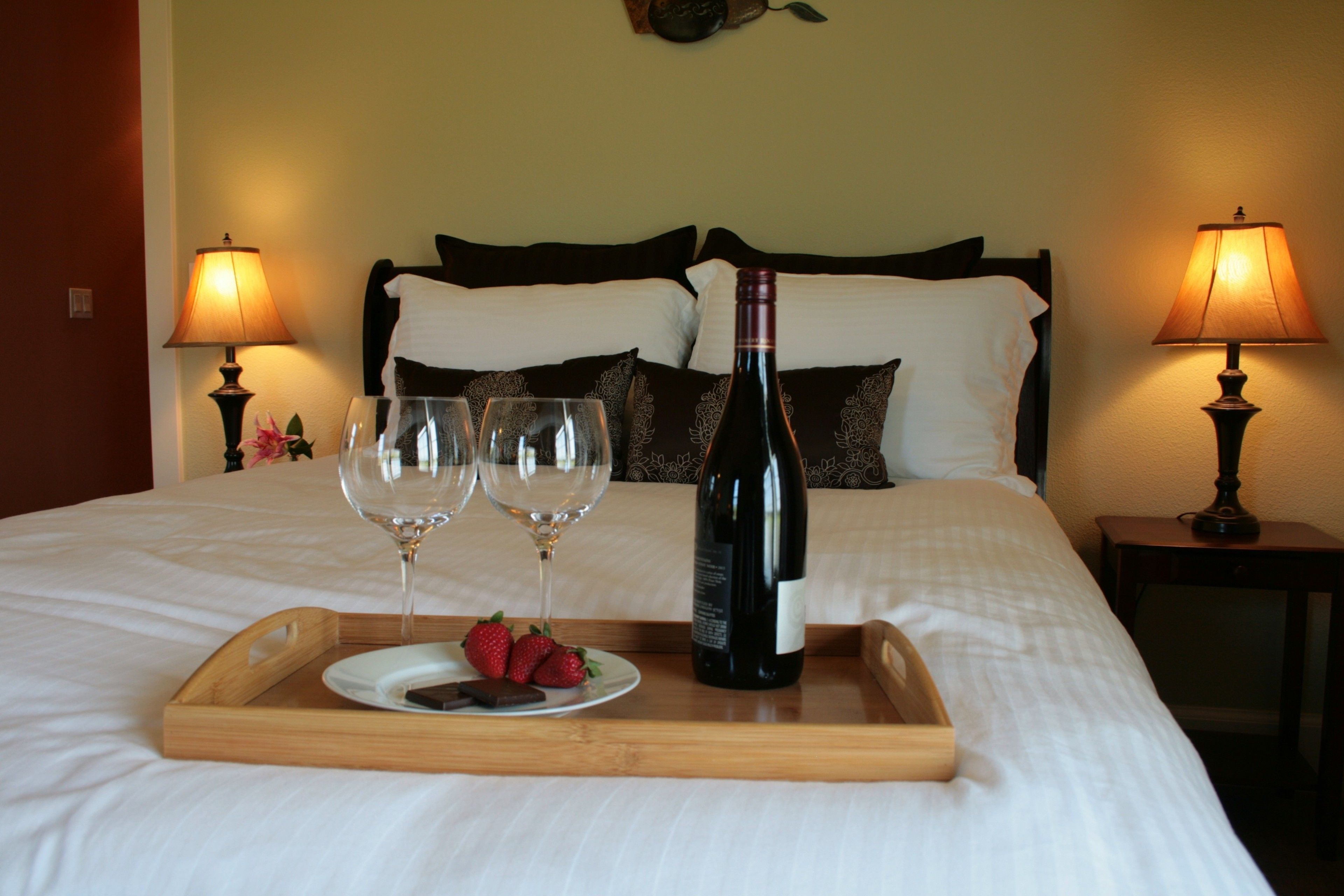 A neatly made bed with a tray of strawberries and two wine glasses, a bottle of wine, and two lamps on either side.