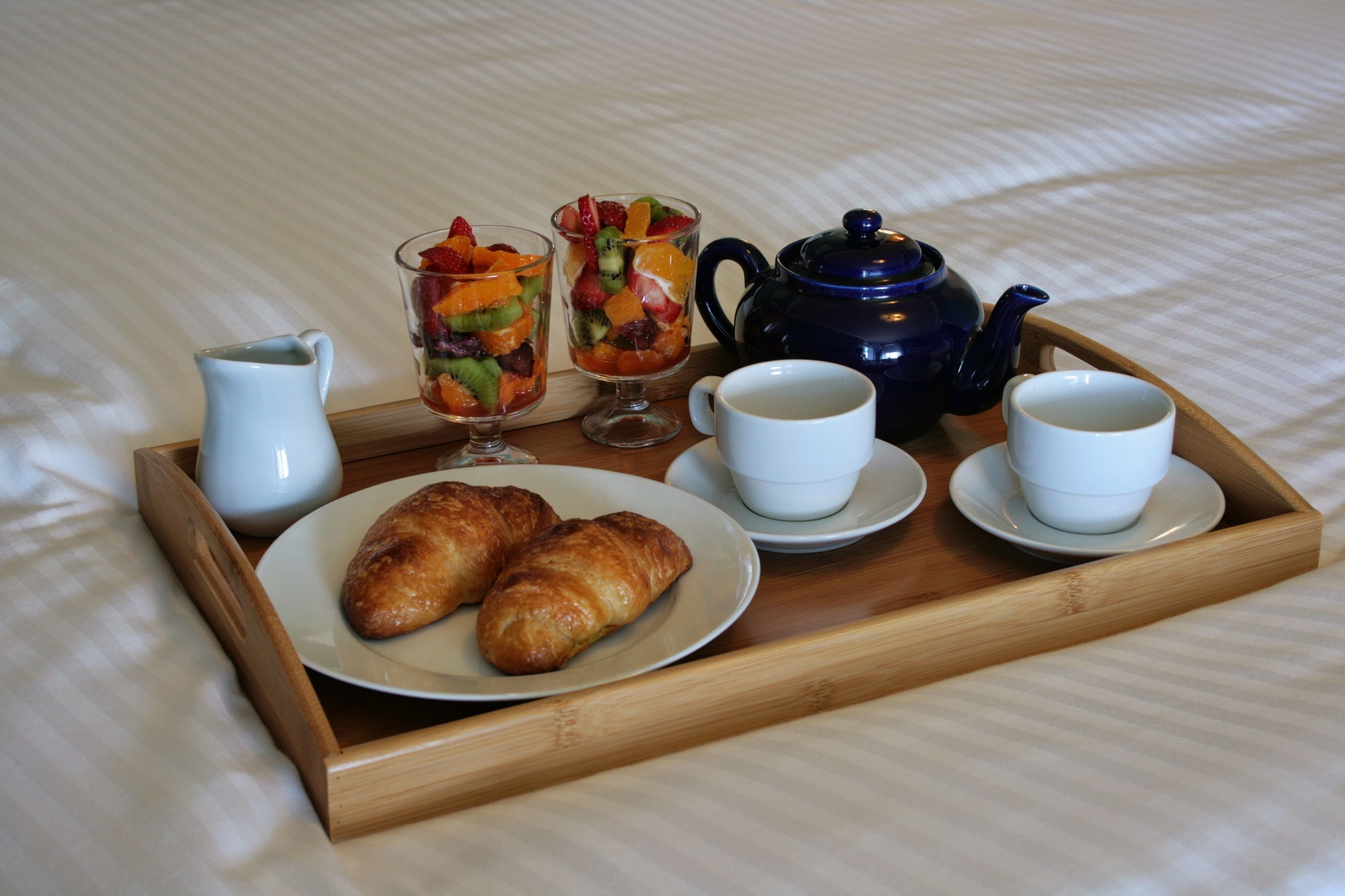 Un plateau contenant des croissants, une salade de fruits, des tasses à café et une théière, posé sur un lit.