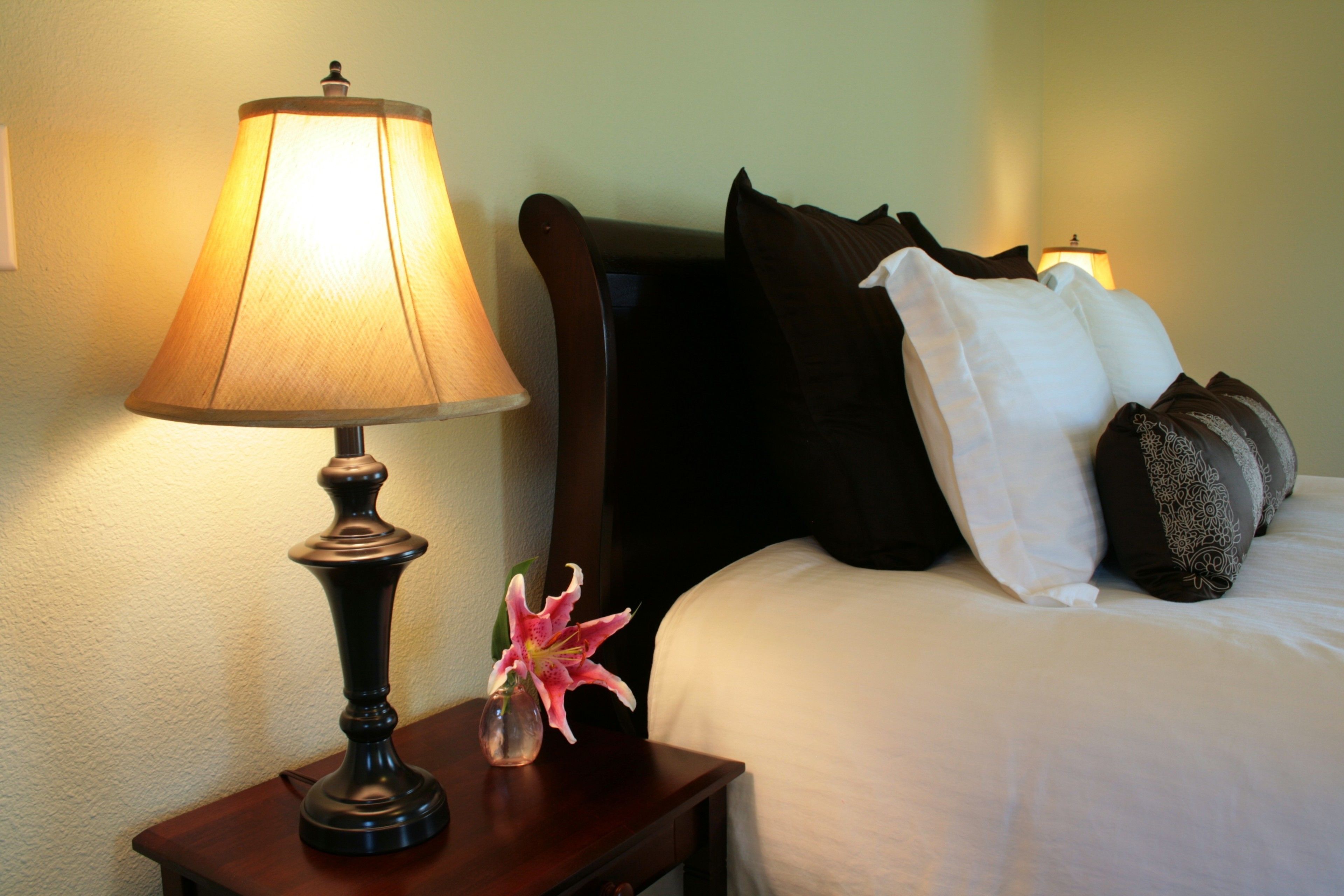 Un lit bien fait, avec des oreillers blancs et à motifs, une lampe de chevet et un vase contenant une fleur rose, posés sur une table de chevet en bois.