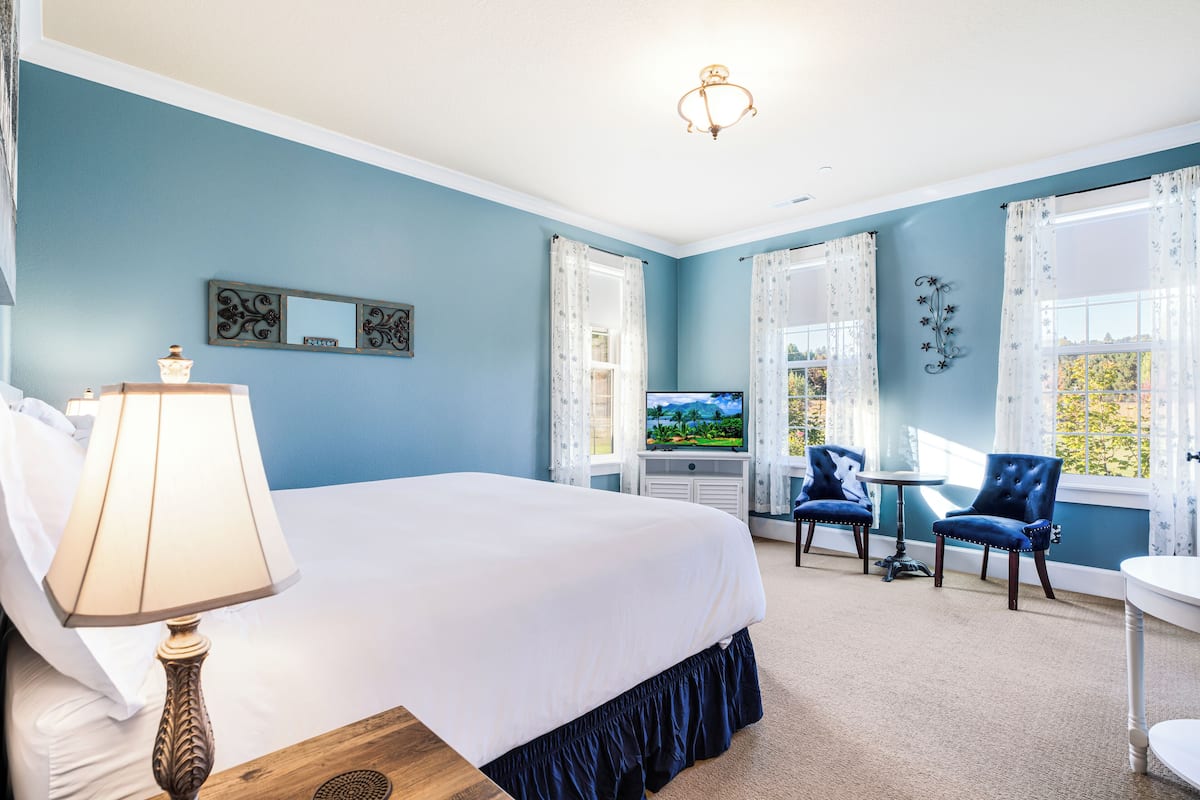 A hotel room with a large bed, a TV, two chairs, a small table, and a window with curtains.