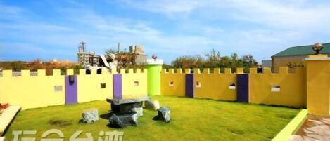 A courtyard with a yellow wall, a green lawn, and a stone structure.