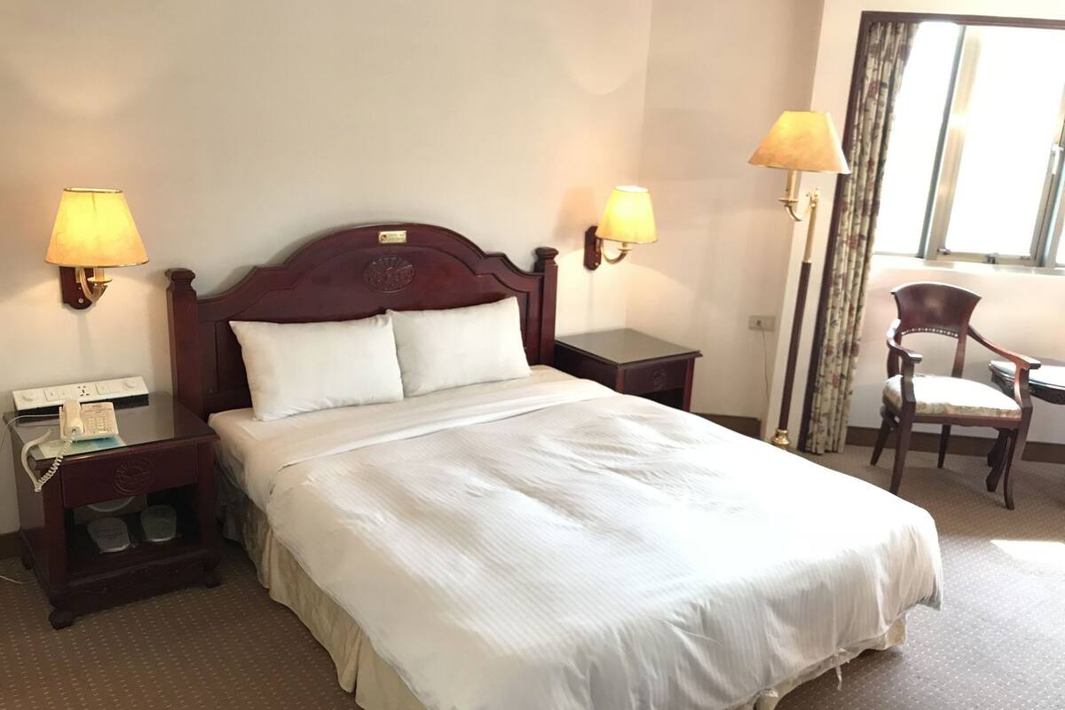 A hotel room with a large bed, two bedside lamps, a chair, a desk, and a window with curtains.