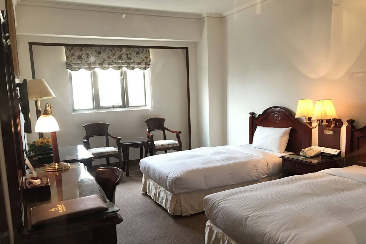 A hotel room with two beds, a desk with a lamp, a chair, a small table, and a window with curtains.