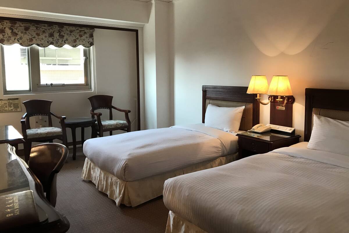 A hotel room with two beds, a desk, and a chair. There is a window with a view of the outside.