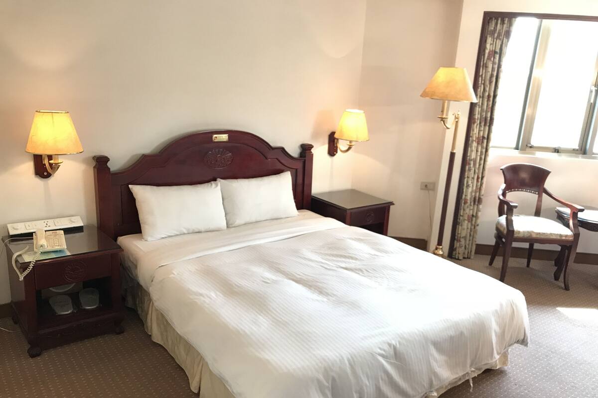 A hotel room with a large bed, two bedside lamps, a nightstand with a phone, a chair, and a window with curtains.
