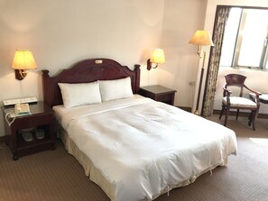 Une chambre d’hôtel avec un grand lit, deux lampes de chevet, une table de chevet avec un téléphone, une chaise et une fenêtre avec des rideaux.