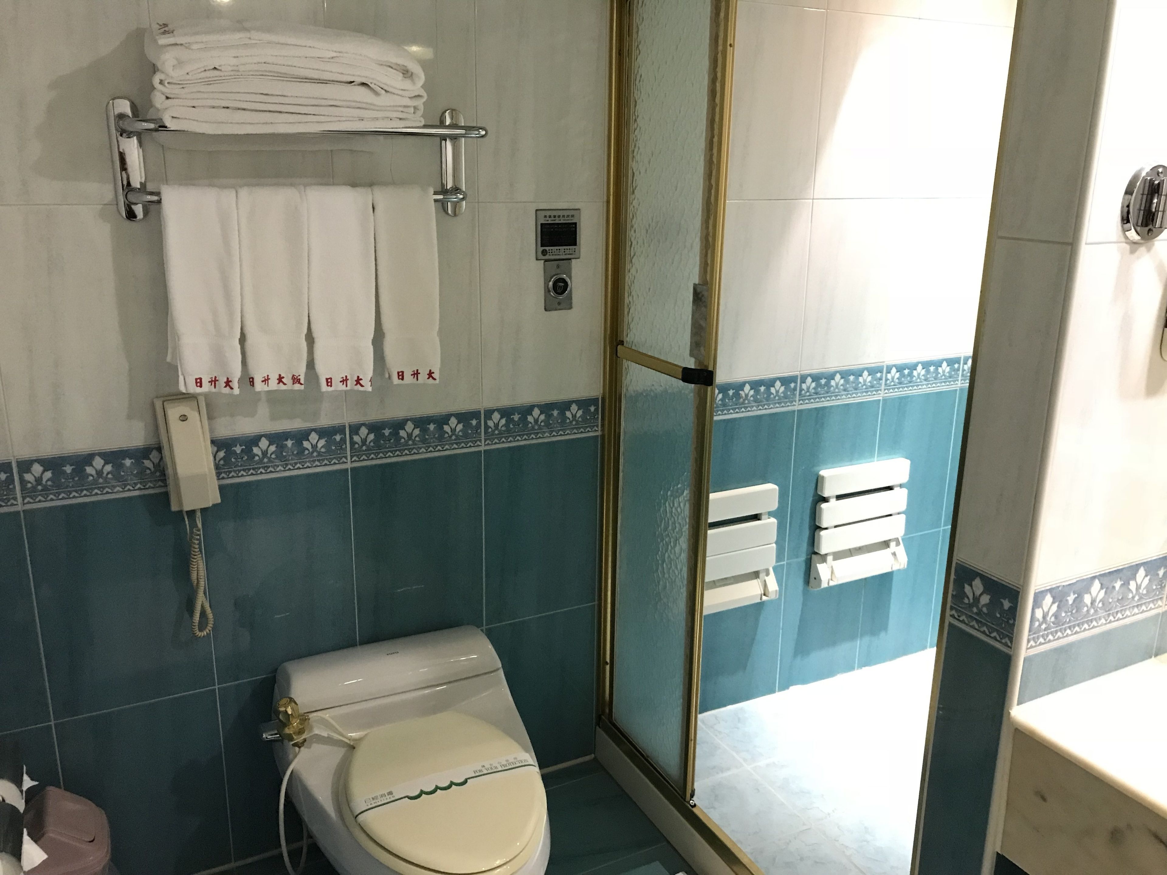A bathroom with a toilet, a towel rack with folded towels, a shower area, and a toilet paper holder.