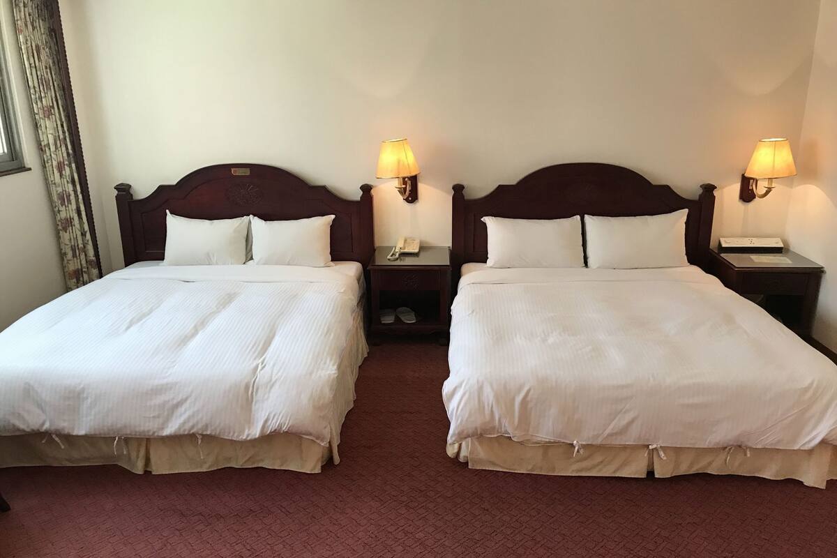 A hotel room with two beds, each with a wooden headboard, white bedding, and bedside lamps.