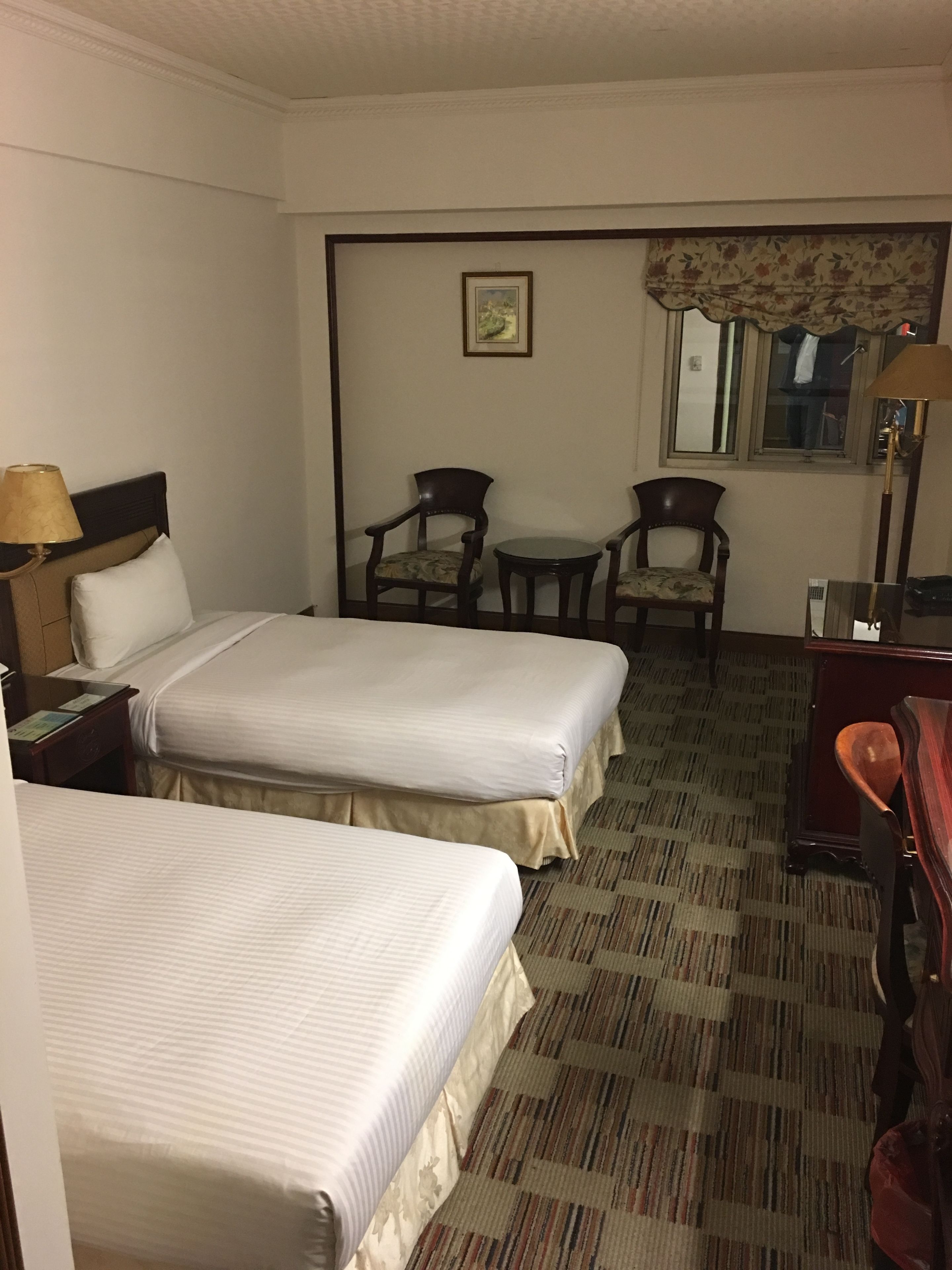 A hotel room with two beds, a desk, a chair, a small table, a lamp, and a window with a curtain.