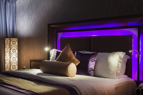 A hotel room with a bed, pillows, a nightstand, and a wall-mounted lamp.