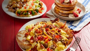 A breakfast spread on a red table with pancakes, scrambled eggs, and hash browns.