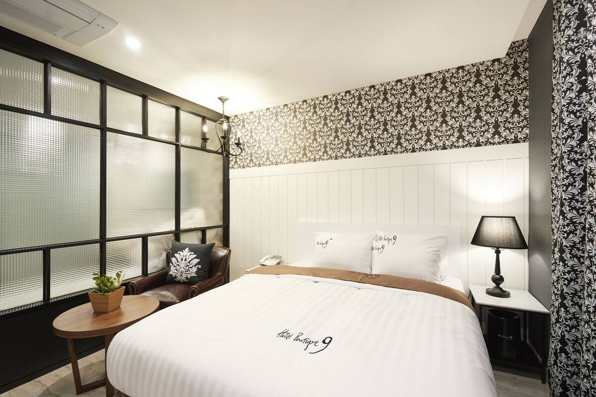 Una habitación de hotel con una cama grande, una mesita con una planta, una silla, una mesita de noche con lámpara y una pared decorativa con estampado floral.