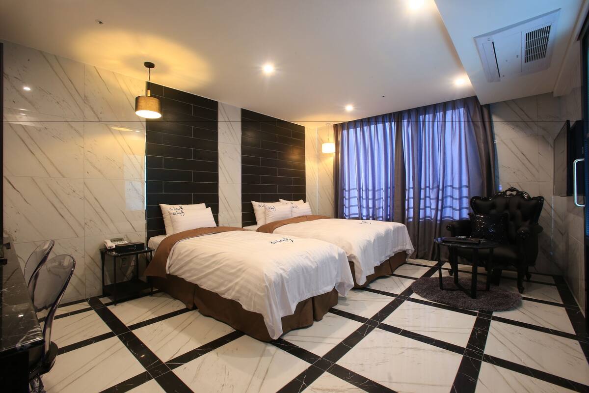 Habitación de hotel con dos camas, zona de estar con una silla, una mesita y un ventanal grande con cortinas.