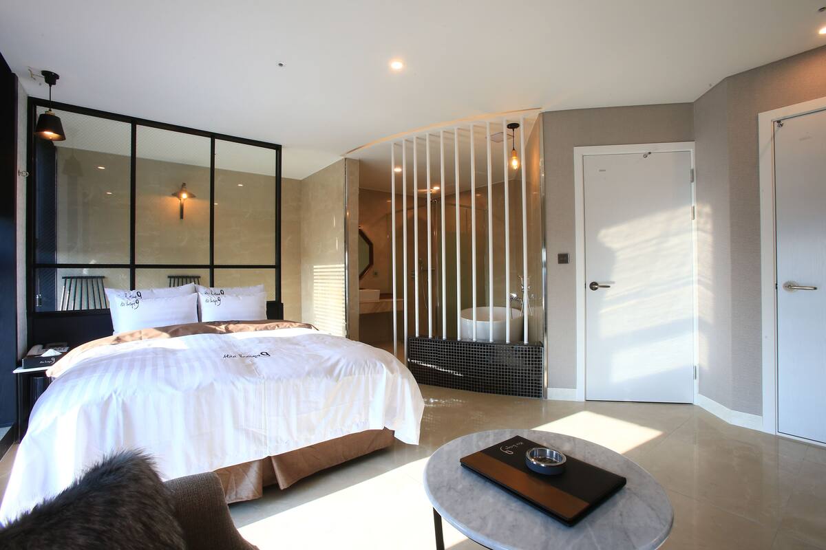 Una habitación de hotel moderna con una cama grande, una mesita redonda pequeña, un baño con bañera y un espejo amplio.