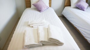 A hotel room with two single beds, each with a white bedspread and a purple pillow. There are folded white towels on the bed in the foreground.
