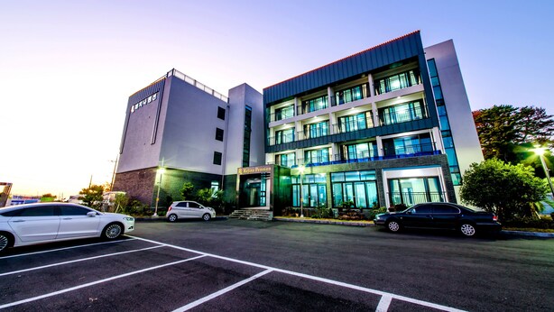A multi-story building with a modern design, featuring a prominent entrance and a parking lot with several cars. - Rejina Pension (Seogwipo)