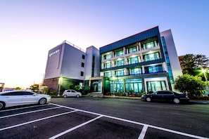 A multi-story building with a modern design, featuring a prominent entrance and a parking lot with several cars. - Rejina Pension (Seogwipo)