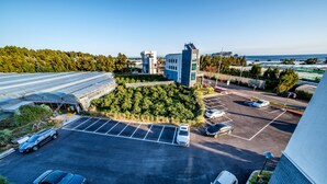 A parking lot with several cars, a building with a flat roof, and a hillside with trees. - Rejina Pension (Seogwipo)