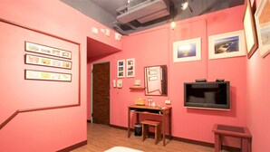 Una habitación en tonos rosa con cuadros enmarcados en las paredes, una mesa de madera con un cesto de basura y un televisor a la derecha.