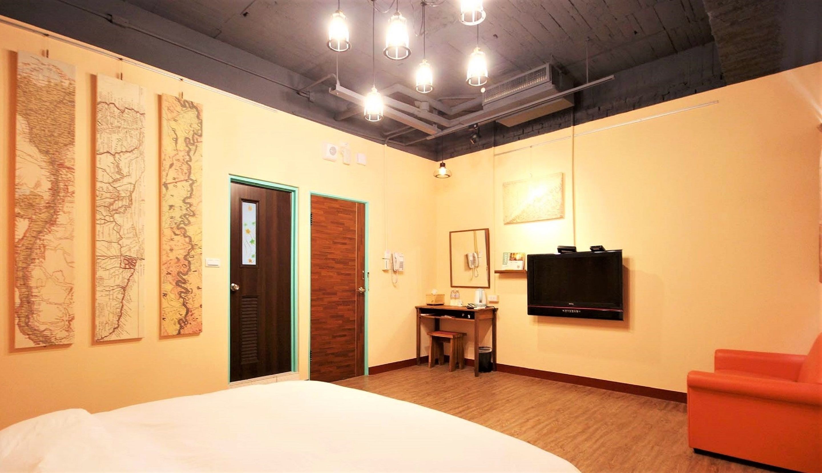Habitación de hotel con cama, escritorio con silla, televisor y tres mapas grandes en la pared.