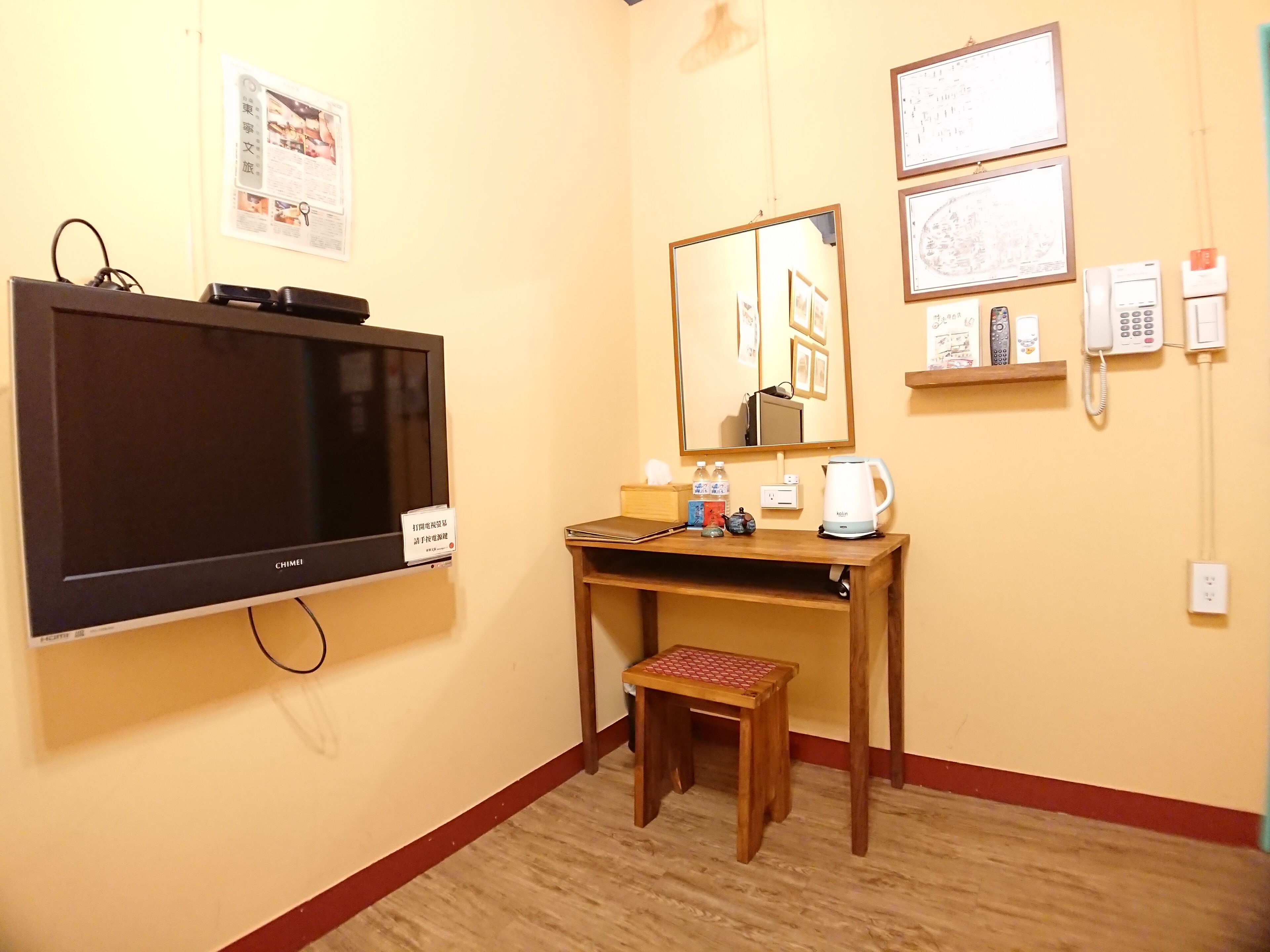 A hotel room with a TV, a wooden desk, a small stool, a mirror, and a wall-mounted phone.