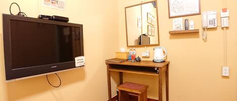 A hotel room with a TV, a wooden desk, a small stool, a mirror, and a wall-mounted phone.