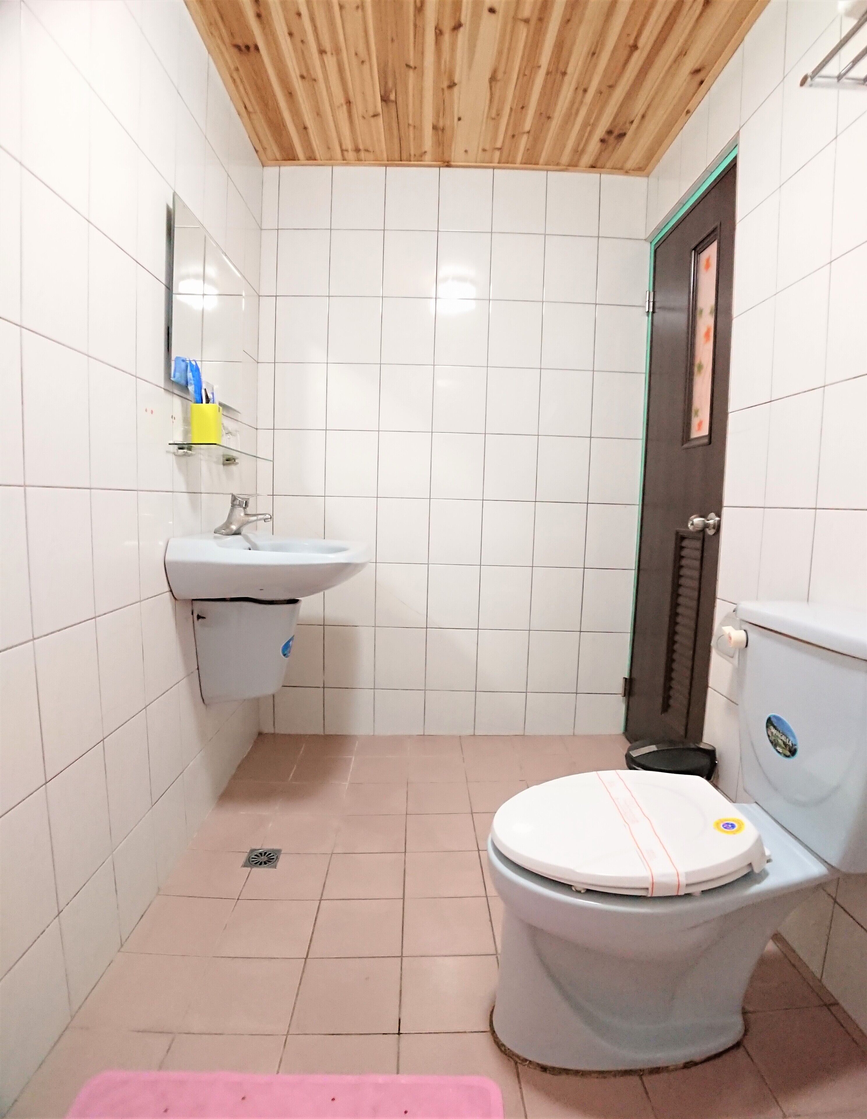 Une petite salle de bain avec des carreaux blancs, un plafond en bois, un lavabo, des toilettes et un tapis rose.