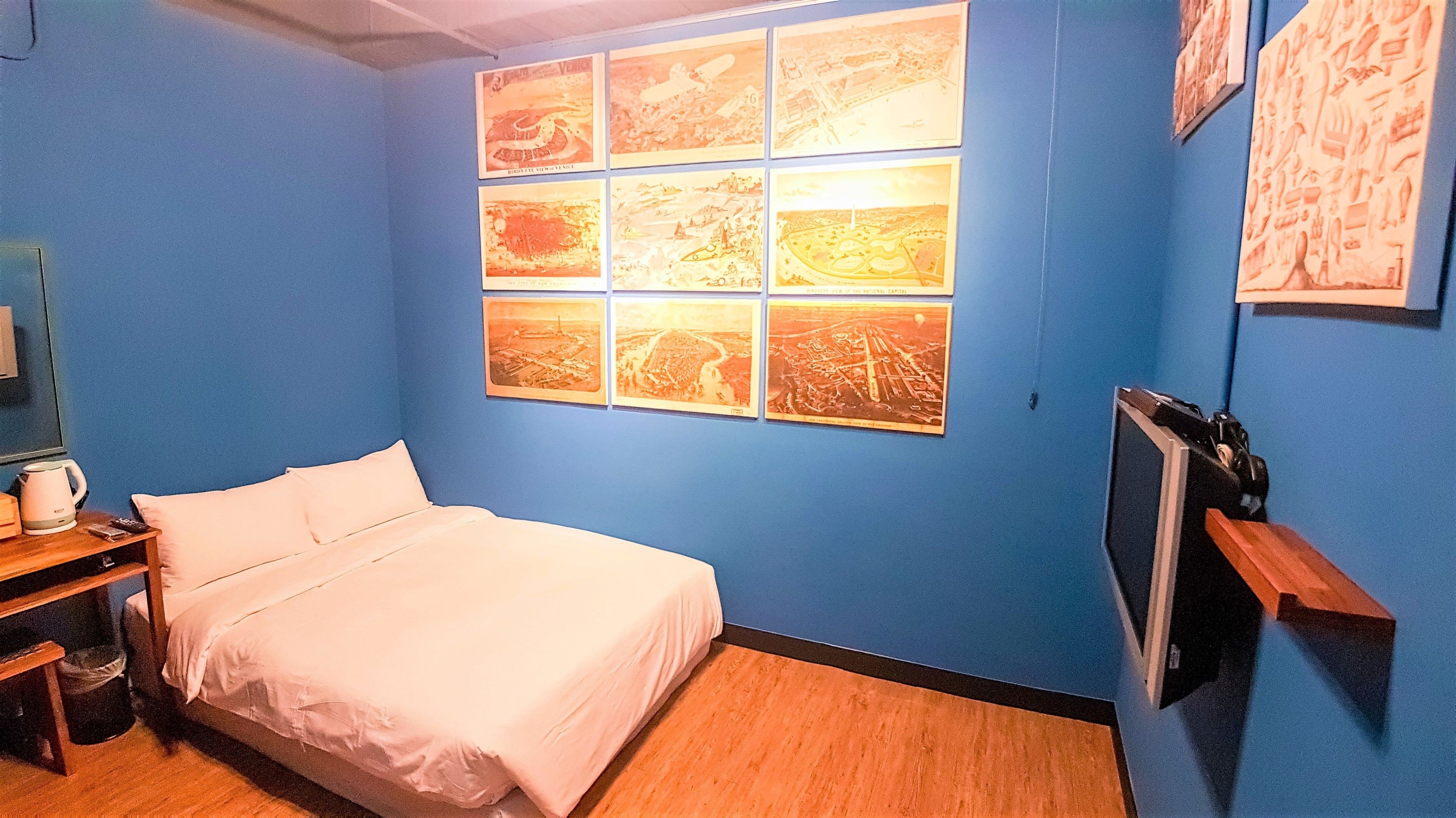 A hotel room with a bed, a desk with a kettle, and a wall with multiple framed pictures.