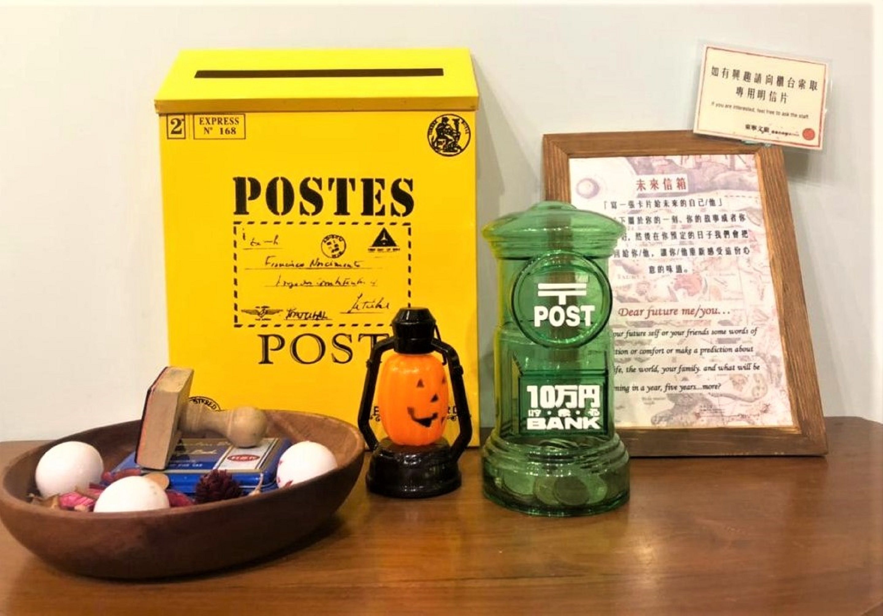 A yellow box labeled 'POSTES' with handwritten notes, a green water bottle with 'POST' and '10,000 yen' on it, a small orange figurine, and a framed document with text.
